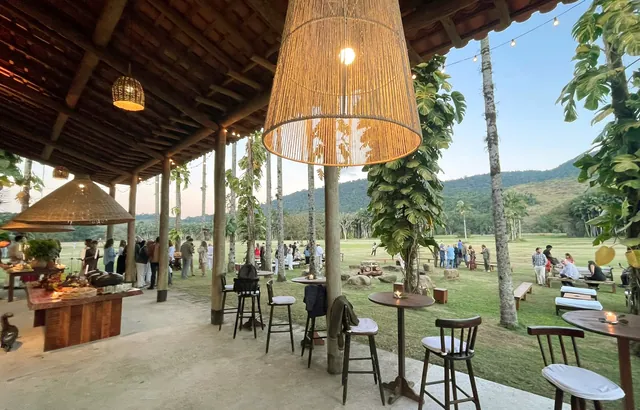 A varanda da casa de apoio ao entardecer: pilares de eucalipto, telhado de madeira e luminárias de palha acesas, com o gramado ao fundo